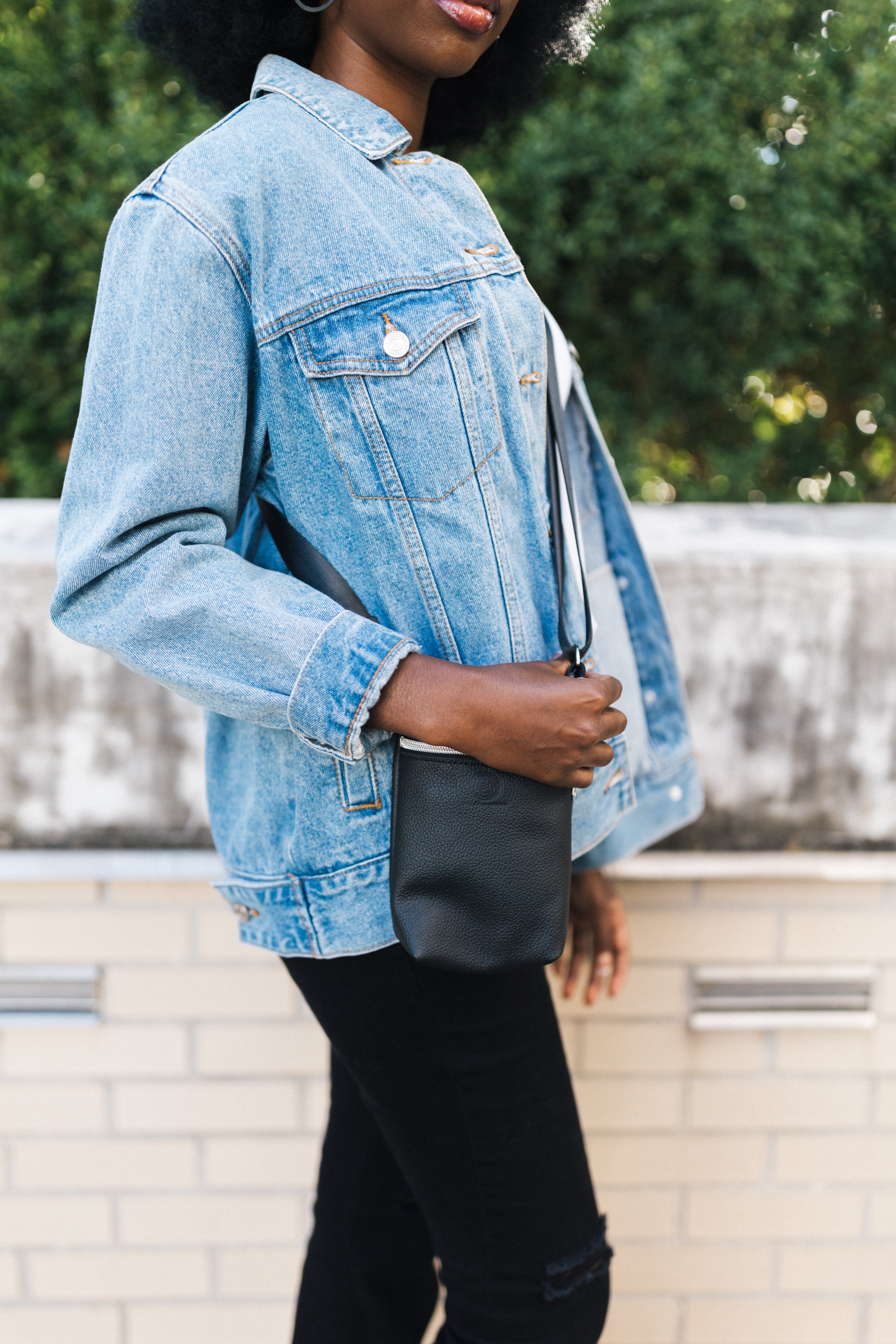 Person wearing a denim jacket holding a leather black crossbody outdoors.