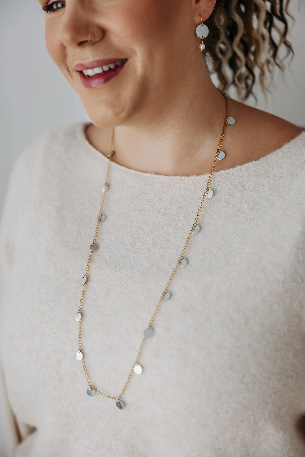 Woman wearing a long gold necklace with small round silver pendants against a neutral background