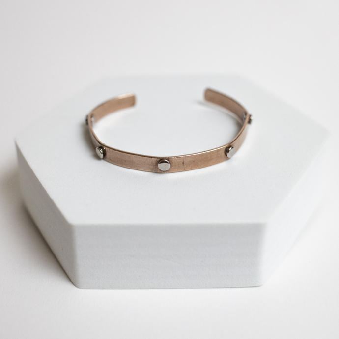 Rose gold bracelet with steel rivets for sale by Mend on the Move on a white hexagonal stand with a white background  
