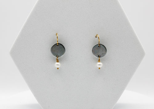 Pair of earrings with round gray steel discs and white pearl beads on a white hexagonal stand.