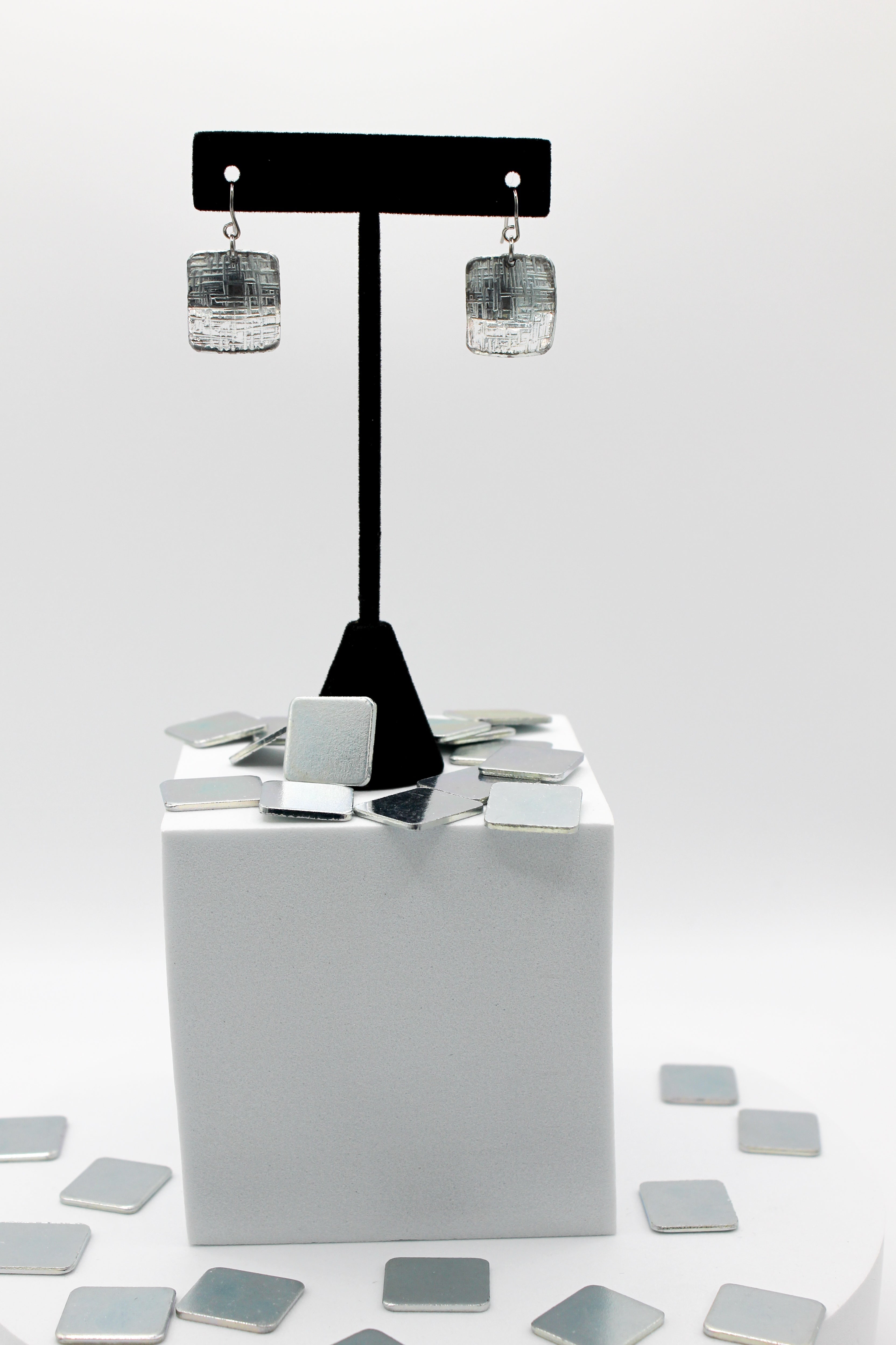 Silver square earrings on a black stand with scattered square mirrors on a white background